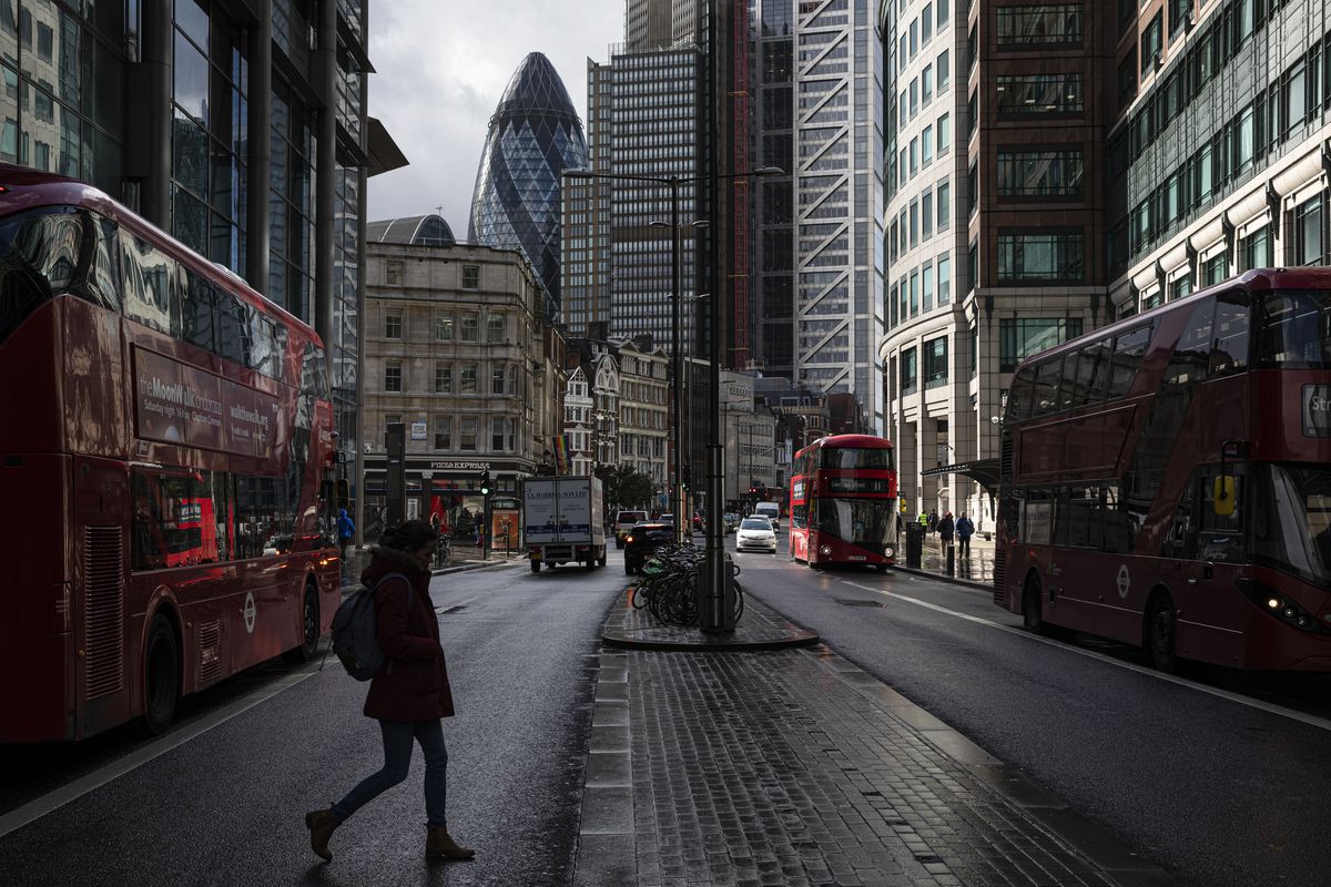 City street with red double-decker buses, tall modern buildings, and a pedestrian crossing under overcast light, in a restrained documentary mood